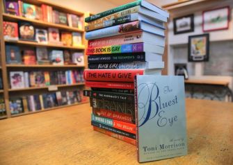 A stack of books in a library setting, including "The Bluest Eye" by Toni Morrison prominently featured in the foreground.