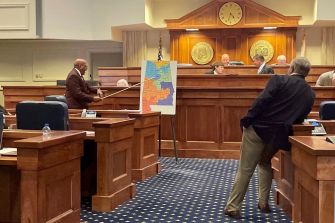 A man points to a map of Alabama's congressional districts during a legislative meeting, with officials seated at a table in the background.