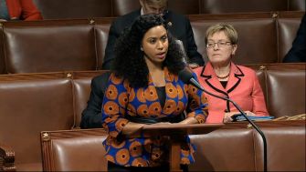 US Rep. Ayanna Pressley speaking at a podium in the House of Representatives, with Senator Tammy Duckworth seated behind her.