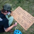 A person sits on the ground writing a message on a sign that reads "Kenosha, WI PD YOUR RACISM IS SHOWING." A person sits on the ground writing a message on a sign that reads "Kenosha, WI PD YOUR RACISM IS SHOWING."