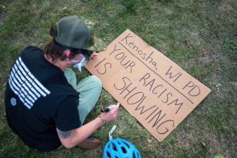A person sits on the ground writing a message on a sign that reads "Kenosha, WI PD YOUR RACISM IS SHOWING."