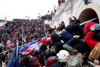 A large crowd of protesters outside the U.S. Capitol building during the January 6 event, with individuals climbing the barriers and waving flags.