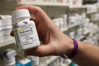 A hand holding a pill bottle of OxyContin, with shelves of other medication in the background.