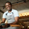 A smiling male restaurant worker in an apron holds cash while sitting at a table. A smiling male restaurant worker in an apron holds cash while sitting at a table.