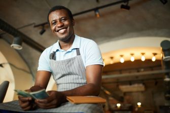 A smiling male restaurant worker in an apron holds cash while sitting at a table.