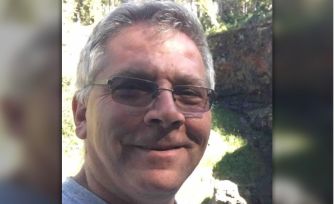 A smiling man with glasses and a gray shirt, standing outdoors with greenery in the background.