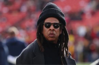 Jay-Z wearing sunglasses and a warm hat at a sporting event.