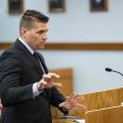 A man in a suit gestures while speaking at a courtroom podium, with a blurred audience visible in the background. A man in a suit gestures while speaking at a courtroom podium, with a blurred audience visible in the background.