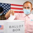 A voter casting a ballot into a ballot box, with another person in the background and an American flag displayed. A voter casting a ballot into a ballot box, with another person in the background and an American flag displayed.