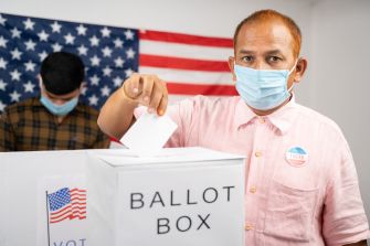 A voter casting a ballot into a ballot box, with another person in the background and an American flag displayed.