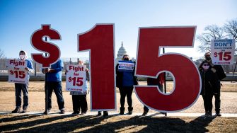 Group of people holding signs advocating for a $15 minimum wage beside a large "$15" display.