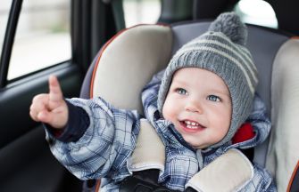 A smiling toddler in a winter coat and knitted hat sits in a car seat, pointing with enthusiasm.