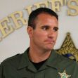 A sheriff in uniform standing in front of the Pasco County Sheriff's Office emblem. A sheriff in uniform standing in front of the Pasco County Sheriff's Office emblem.