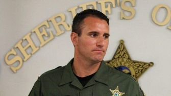 A sheriff in uniform standing in front of the Pasco County Sheriff's Office emblem.