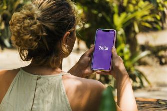 A woman holding a smartphone displaying the Zelle app logo, surrounded by greenery.