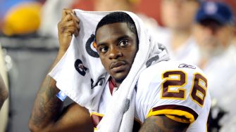 Former NFL player sitting on the sidelines with a towel over his head, wearing a jersey.