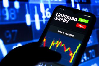 A close-up of a smartphone displaying the Goldman Sachs stock trading app, with stock market data in the background.