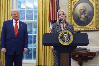 A woman speaking at a podium with the presidential seal and a man standing nearby in an official setting, with historical portraits visible in the background.