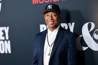Russell Simmons smiling at a public event, wearing a baseball cap and suit.