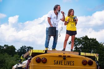 A man and woman standing on top of a school bus with a speaker and a sign that reads "SAVE LIVES," advocating for gun reform.