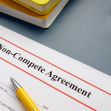 A close-up of a non-compete agreement document on a desk, accompanied by a yellow pen and reading glasses. A close-up of a non-compete agreement document on a desk, accompanied by a yellow pen and reading glasses.