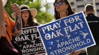 Two women holding signs supporting the protection of Oak Flat, with one sign stating "Pray for Oak Flat" and the other reading "Protect Oak Flat."