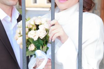 A woman in a white coat holding a bouquet of roses, standing behind metal bars, with a man in a suit beside her.