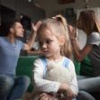 A young girl holding a teddy bear looks worried as her parents argue in the background. A young girl holding a teddy bear looks worried as her parents argue in the background.