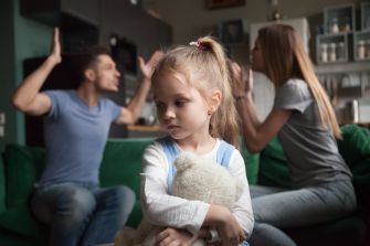 A young girl holding a teddy bear looks worried as her parents argue in the background.