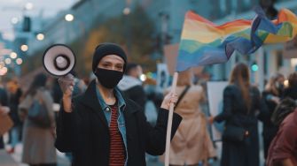 A person holding a megaphone and a rainbow flag at a rally, surrounded by a crowd.