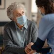 An elderly woman and a caregiver wearing masks engage in a meaningful conversation in a nursing home setting. An elderly woman and a caregiver wearing masks engage in a meaningful conversation in a nursing home setting.