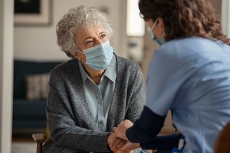 An elderly woman and a caregiver wearing masks engage in a meaningful conversation in a nursing home setting.