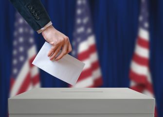 A person casting a ballot into a voting box, with American flags in the background.