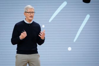 Tim Cook speaking at a presentation event with a visually abstract background.