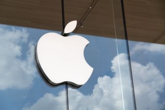 Apple logo displayed on a glass storefront with clouds reflecting in the background.