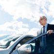A man in a suit standing next to a silver car with a partly cloudy sky in the background. A man in a suit standing next to a silver car with a partly cloudy sky in the background.