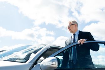 A man in a suit standing next to a silver car with a partly cloudy sky in the background.