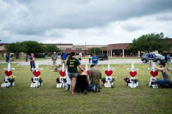 A group of people memorializing victims of the Santa Fe High School shooting, with crosses and flowers displayed in a field outside the school building.
