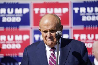 Rudy Giuliani speaking at a press conference with Trump campaign signage in the background.