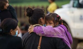 A woman holds a child in an outdoor setting, with others in the background, highlighting the emotional impact of immigration policies.
