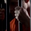 A close-up of a person in an orange prison jumpsuit holding onto prison bars. A close-up of a person in an orange prison jumpsuit holding onto prison bars.