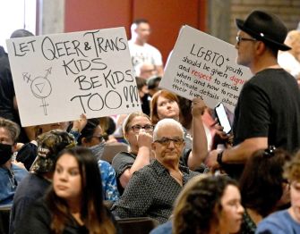 A crowd attending a meeting, with individuals holding signs supporting LGBTQ+ rights and advocating for the protection of queer and trans youth.
