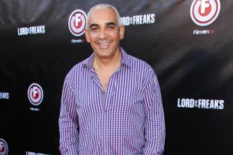 A man posing for a photo at a promotional event with a backdrop featuring the title "Lord of Freaks" and the logo for FilmOn TV.