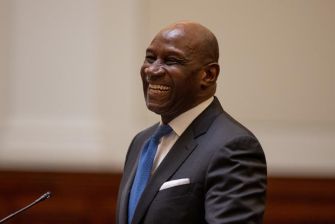 A smiling man in a dark suit and blue tie stands in a formal setting, likely during a ceremony.