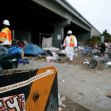 Construction workers in safety gear near a homeless encampment under a freeway, with debris and tents visible. Construction workers in safety gear near a homeless encampment under a freeway, with debris and tents visible.