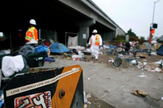 Construction workers in safety gear near a homeless encampment under a freeway, with debris and tents visible.