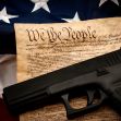 A handgun resting on a historical document with the text "We The People," symbolizing the intersection of gun rights and constitutional law, alongside an American flag. A handgun resting on a historical document with the text "We The People," symbolizing the intersection of gun rights and constitutional law, alongside an American flag.