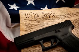 A handgun resting on a historical document with the text "We The People," symbolizing the intersection of gun rights and constitutional law, alongside an American flag.