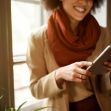A person standing near a window, smiling while using a tablet, with plants visible in the foreground. A person standing near a window, smiling while using a tablet, with plants visible in the foreground.