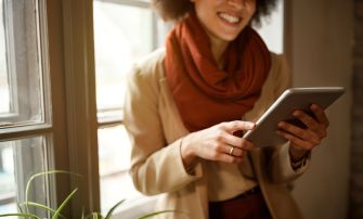 A person standing near a window, smiling while using a tablet, with plants visible in the foreground.